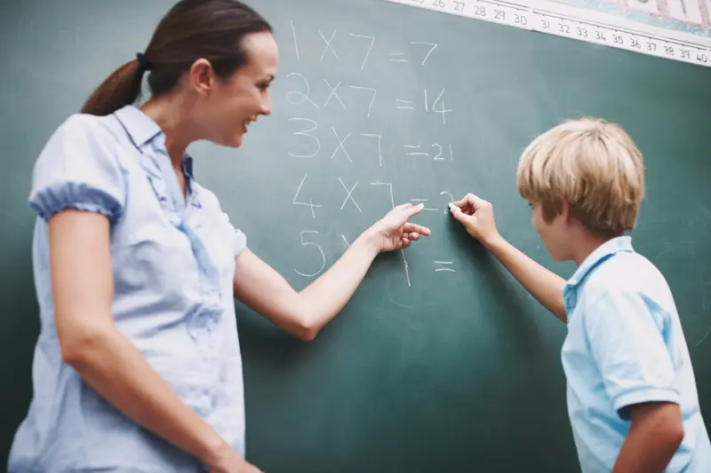 Profesoară explicând înmulțiri la tablă, alături de un elev
