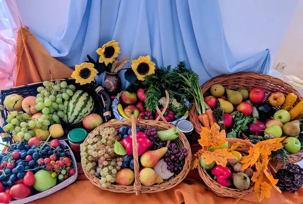 Aranjament de toamnă cu coșuri pline de fructe și legume, floarea-soarelui și frunze decorative pe un fundal de draperii albastre.