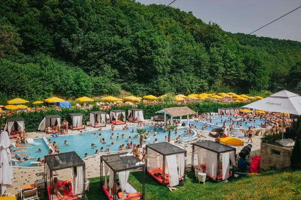 Ștrandul Sun Tropez vara: piscine cu apă albastră, baldachine, umbrele galbene și pădure pe deal în spate